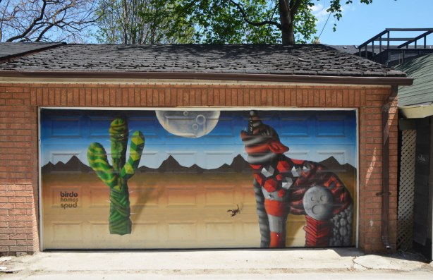 street art mural on a garage door in an alley by birdo - a coyote howling at the moon in the desert with a large cactus in the picture as well as a scorpion. words: birdo homes spud