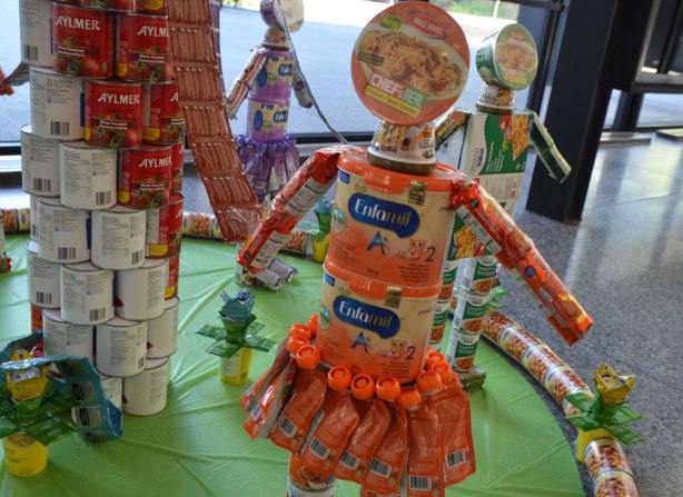models of children made with packaged food. A girl is made with orange enfamil containers, bowl of noodles for head, tubes with orange packaging as a skirt, and cyclindrical packages of cookies as arms