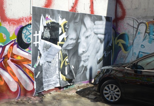 a large black and white photojournalist picture, part of CONTACT photography festival, pasted on a wall - an ill Chinese man is being comforted by his wife.  The picture is on a graffiti covered wall and a black car is parked close to it. 