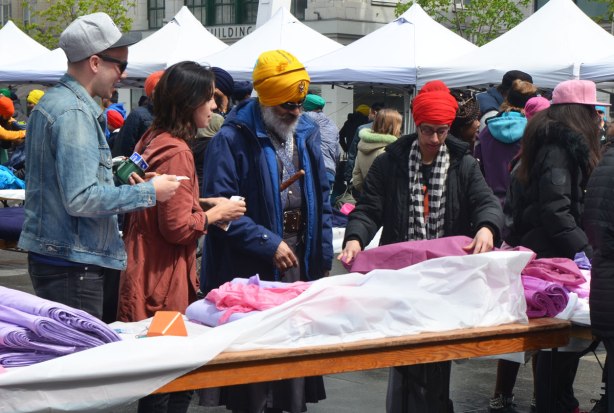 People having their heads wrapped in a turban, many different colours, at an event at Yonge Dundas square run by the Sikh Youth Federation. Sikh volunteers are making the turbans using stacks of fabric laid out on 5 long tables. Choosing magenta fabric