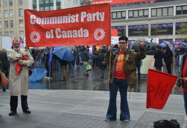 An older man holds one end of a red banner for the Communist Party of Canada while a young man holds the other end - May day, International Workers Day rally at Dundas Square on a rainy day - 