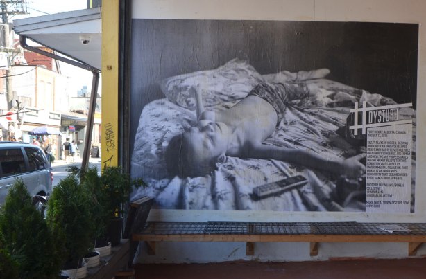 a large black and white photojournalist picture, part of CONTACT photography festival, pasted on a wall - a sick boy is lying on a bed, on a wall in the entranceway to a store, sidewalk and street scene in the background