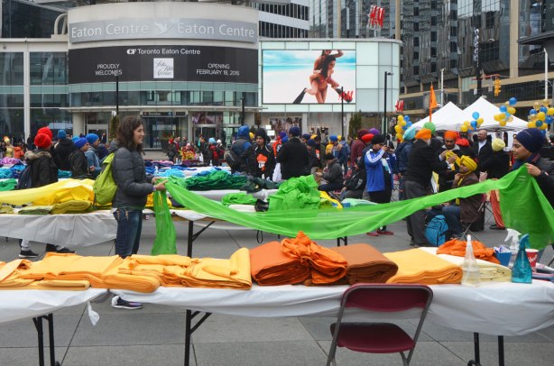 tables with piles of colourful fabric outside at Yonge Dundas square with the signs and billboards of the Eaton Centre behind. Many people are in the square. 