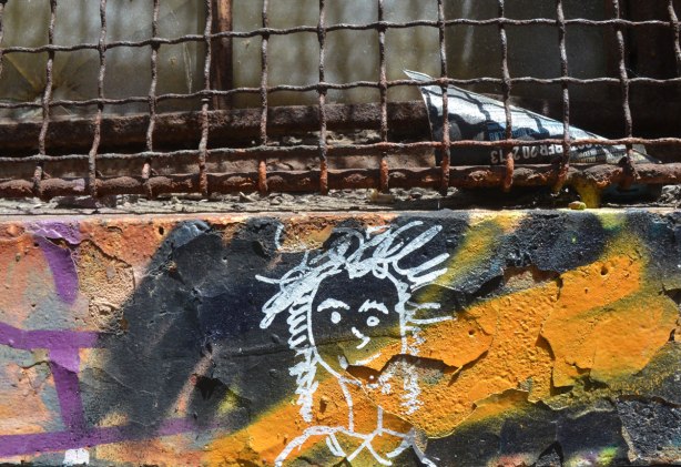 under the rusted metal of a wire grill over a window, on a wall that was painted in black and orange street art, there is a small white line drawing of a girl's head