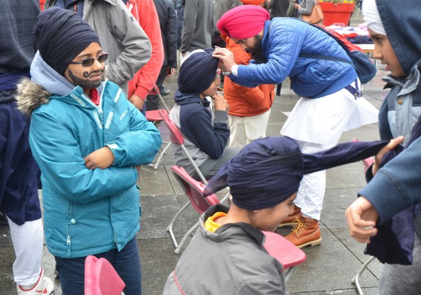 People having their heads wrapped in a turban, many different colours, at an event at Yonge Dundas square run by the Sikh Youth Federation. Sikh volunteers are making the turbans using stacks of fabric laid out on 5 long tables. A boy with a black turban and his face painted with a black beard and mustache watches others get turbans