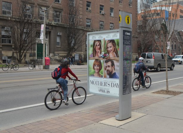 cyclists ride by an information pillar that has a large ad jutting out towards the street. 