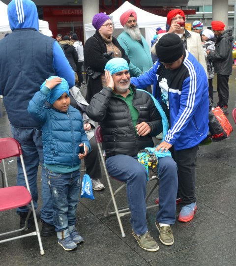 People having their heads wrapped in a turban, many different colours, at an event at Yonge Dundas square run by the Sikh Youth Federation. Sikh volunteers are making the turbans using stacks of fabric laid out on 5 long tables. A father and son with matching light blue turbans