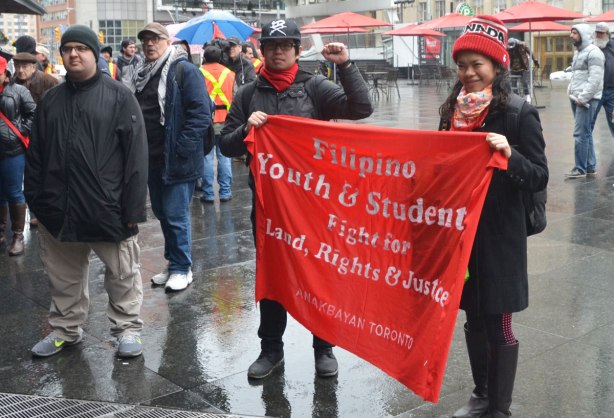 Two students hold a red banner for the Filipino Youth and Students fight for land, rights and justice - May day, International Workers Day rally at Dundas Square on a rainy day - 