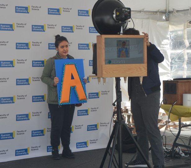 A young woman has her picture taken with a large orange letter A on a blue square. 