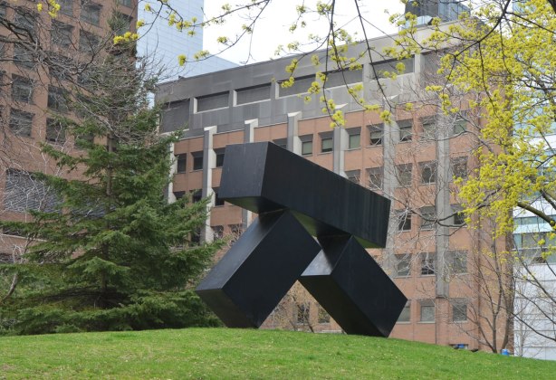 black metal sculpture of three boxes precariously balanced, the upper box is almost horizontal and it is being supported by the other two who are falling down but still balancing 