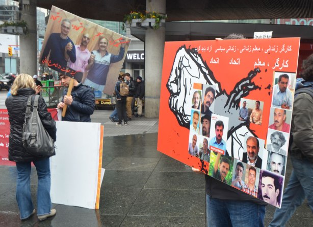 A man holds up a sign written in Arabic about men missing I think. It is red with a picture of a fist on it. On the fist are glued pictures of 16 men . Also in the picture, a man holding a placard with a photo of three men on it, talks to a woman whose back is to the camera. 