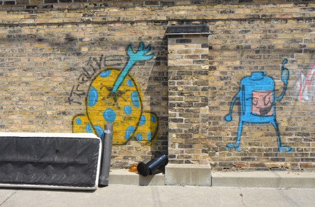 an old mattress leans against an old brick wall that has two pieces of graffiti on it. One is a blue and yellow egg shape with a blue arm and hand sticking out of a crack in it. The words "It's alive" are written beside it. The other is a small blue creature like shape 