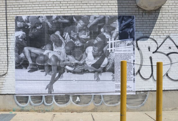 a large black and white photojournalist picture, part of CONTACT photography festival, pasted on a wall - a boat load of Libyan migrants is being rescued from their rubber dinghy. 
