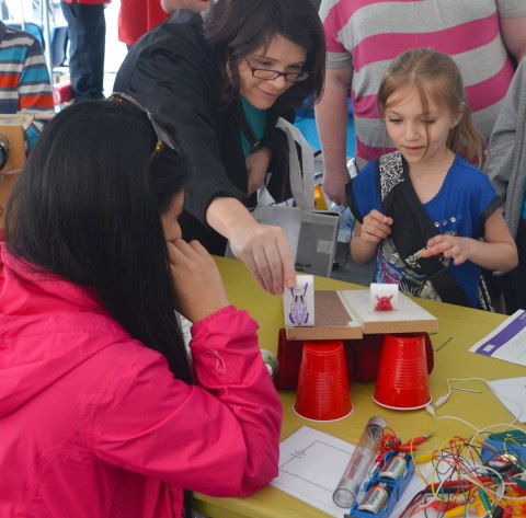 A woman and a girl are racing objects that they made. Inside small rectangular boxes are marbles that make the boxes tumble down an incline. 
