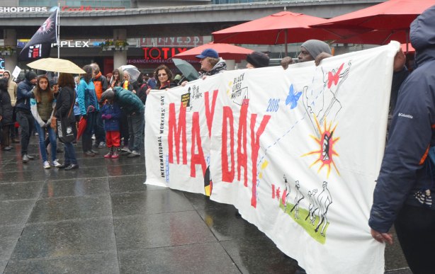 A group of women standing behind and holding up a large white banner for May Day - May day, International Workers Day rally at Dundas Square on a rainy day - 