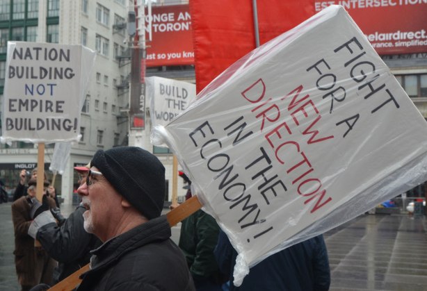A man holds a sign that says "Time for a new direction in the economy" - May day, International Workers Day rally at Dundas Square on a rainy day - 