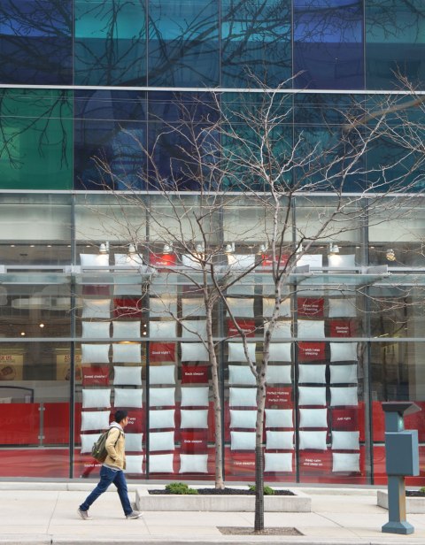 green and blue glass of a building's facade, with a storefront below. The windows of the store are filled with red and white pillows arranged in a grid. 