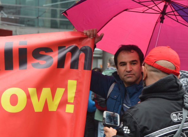 A man under a pink umbrella talks to a man whose back is to the camera - May day, International Workers Day rally at Dundas Square on a rainy day - 