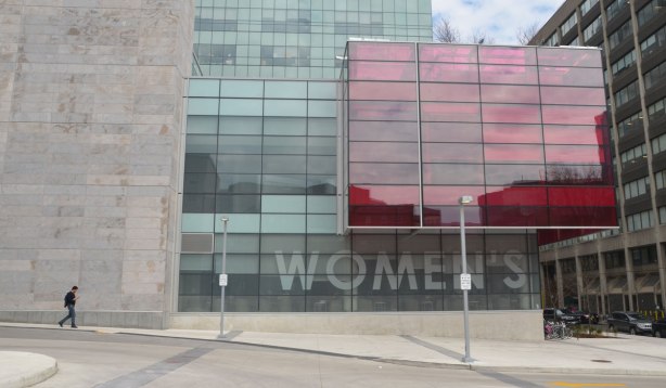 a lone man walks by the new Womens College Hospital building with its light grey stone facade, large glass section, and large pink glass section. 
