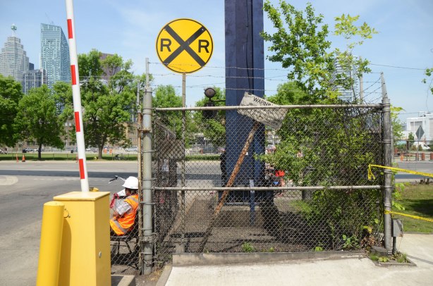 a wire fence with barbed wire across the top, behind the fence is round yellow railway crossing sign as well as a large blue metal pole and a small shrub. There is also an old warning sign for a railway that once ran past here. 