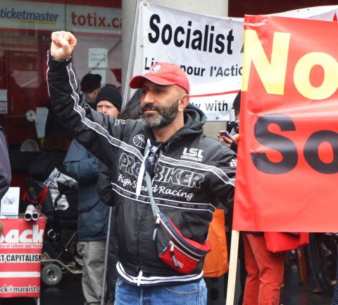 May day, International Workers Day rally at Dundas Square on a rainy day - a man holds his fist in the air as he stands beside a banner 