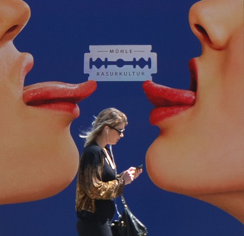 A woman with her phone in her hands walks past a large poster of two women face to face where all you can see is their nose, mouth, chin and tongues that are stuck out. A razor blade balances between the two tongues. 