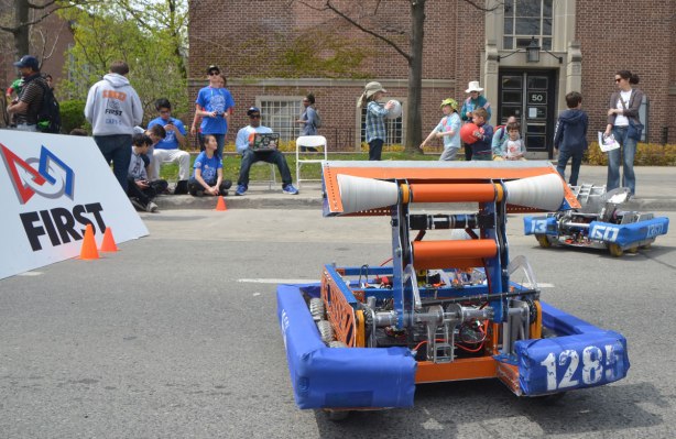 A group of students is sitting on a sidewalk. One of them has a laptop and he is controlling a robot machine with wheels that is moving around on the street. 