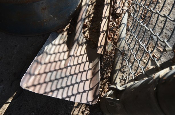 the end of a shovel is in the ground, behind a chainlink fence. The sun is shining and making reflections. The reflection of the chainlink fence is on the shovel. 