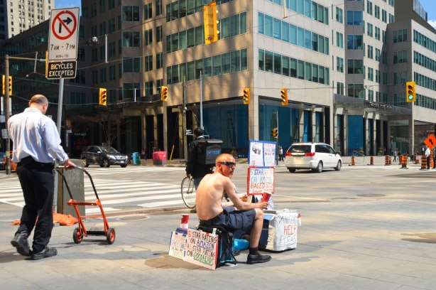 a shirtless man in sitting on a stool at the corner of Front and Union streets. He has four signs (behind, above, in front, and beside him) asking for spare change or for you to buy his novel that is well rated on both Amazon and Goodreads. 