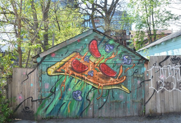 An old wood garage in an alley that is painted by Shalak, a slice of pizza with pepperoni pieces flying off it (or onto it!)