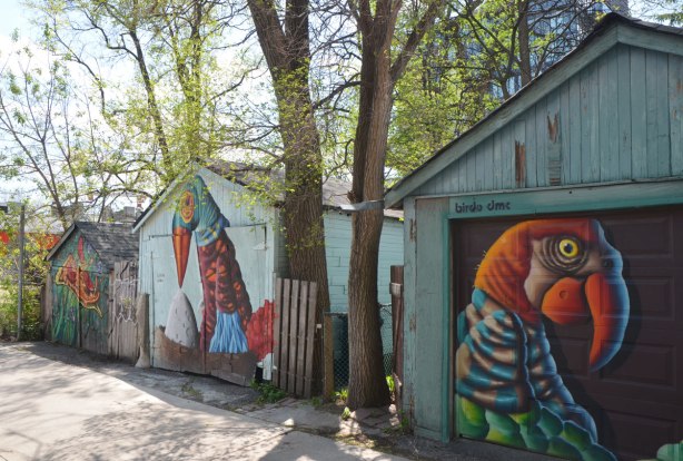 Three garages in an alley, with trees growing between them. The first two have garage doors with birdo murals on them and the third has a pizza street art painting by Shalak 