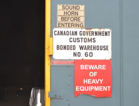 three warning signs on the outside of a door of the Redpath Sugar shed, a warehouse for storing raw sugar. One says "Beware of Heavy Equipment", the second says "Sound horn before entering" and the third says "Canadian Government Customs Bonded Warehouse no. 60"
