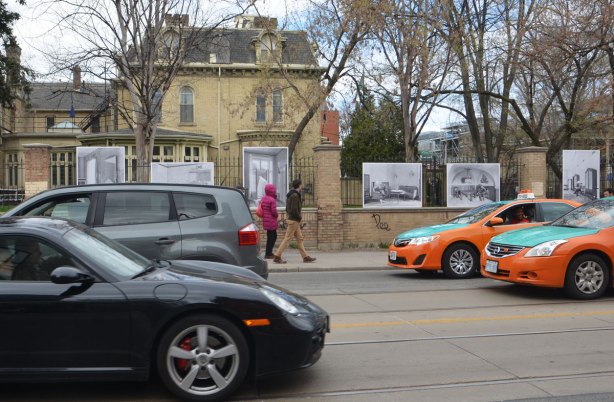 photos mounted along the exterior of a wrought iron fence around the Italian Consulate, right beside the sidewalk. The consulate is on old brick house (mansion) from the 1800's - as seen from across the street with pedestrians walking past and cars driving past 