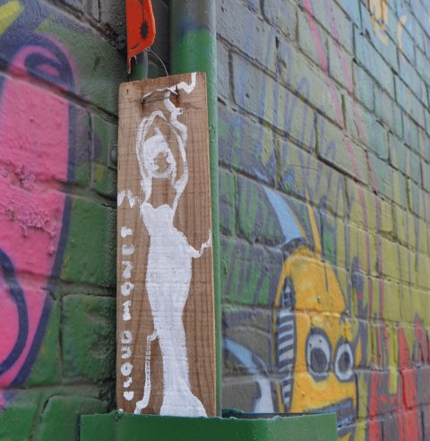 small vertical drawing of a woman, drawn in white, wearing a long form fitting dress, standing, on brown, tied to a wall with a mural laready painted on it. 