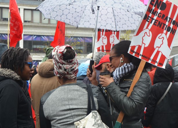 women talking and laughing together at a rally, holding placards that say "End Immigration Detention " and also holding umbrellas
