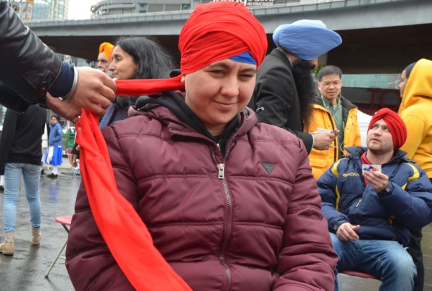 People having their heads wrapped in a turban, many different colours, at an event at Yonge Dundas square run by the Sikh Youth Federation. Sikh volunteers are making the turbans using stacks of fabric laid out on 5 long tables. 