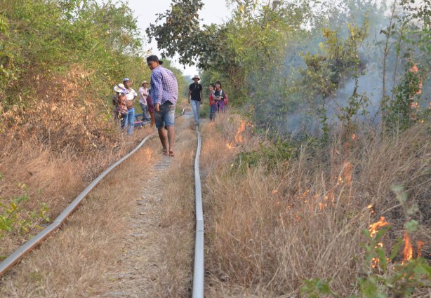 people walking along old train tracks, small fires burn in the bush beside the tracks. 