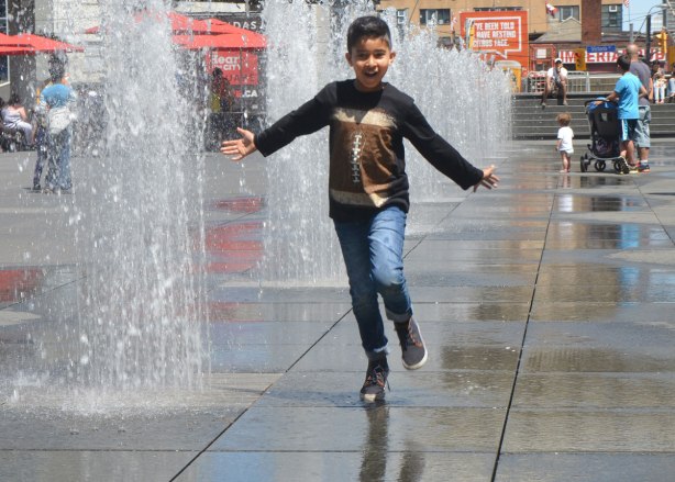 a boy runs down the middle of the fountains at Dundas Square with his arms out at his side and a smile on his face