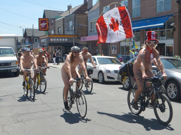 Five or six men on bicycles on a street in Kensington, they are all naked. One is wearing a large Canada Flag hat and has a Canadian flag flying from the back of his bike. Participants in the World Naked Bike Day.