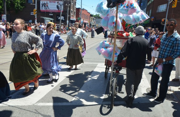 Women dancers in traditional Portuguese dress, dancing in a parade. One of them pauses to look at the man selling cotton candy and popcorn froma cart.
