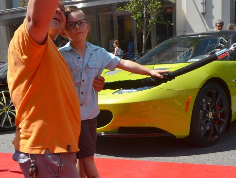 A father is taking a selfie of himself with his son in front of a yellowish green sports car at a car show. 