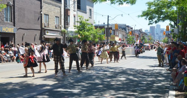 On a street, Dundas West in Toronto, there is a parade, people on the sidewalk watching, and dancers performing in the parade. Portugal Day parade.