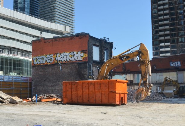 view of a construction site where one old building remains, a digger is on the site as is a very large orange rubbish bin
