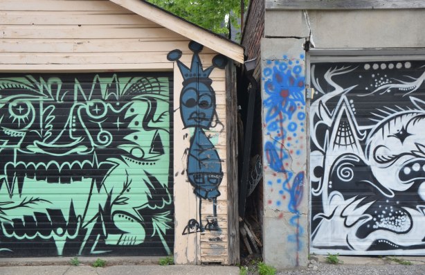 a skinny blue graffiti figure on the edge of a garage, between two garage doors with stylized and abstract faces painted on them. One is black and green and the other is black and white