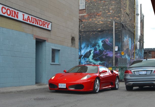 a red Ferrari and a beige Mercedes drive past each other beside a coin laundry in a lane. 