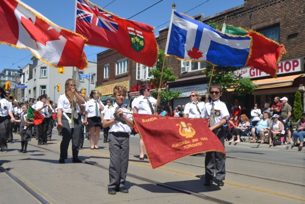 Two boys holding a red banner for a marching band in a parade. Lots of flag holders behind them, a Canadian flag, an Ontario flag, a Toronto flag and a Portuguese flag.