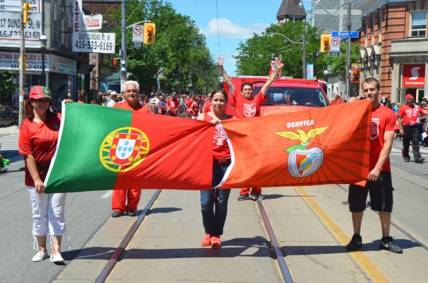 People in a parade, three people holding two flags, Portugal and Benfica. A man behind them is holding his arms up in the air.