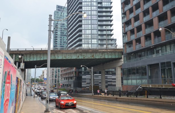 some traffic on Bathurst street on a rainy day, as the street passes under the Gardiner Expressway, condos on the right, construction hoardings on the left. 