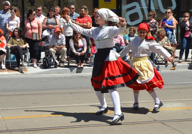 Two girls in traditional Portuguese dresses are dancing in a parade, onlookers sitting on the sidewalk behind them.