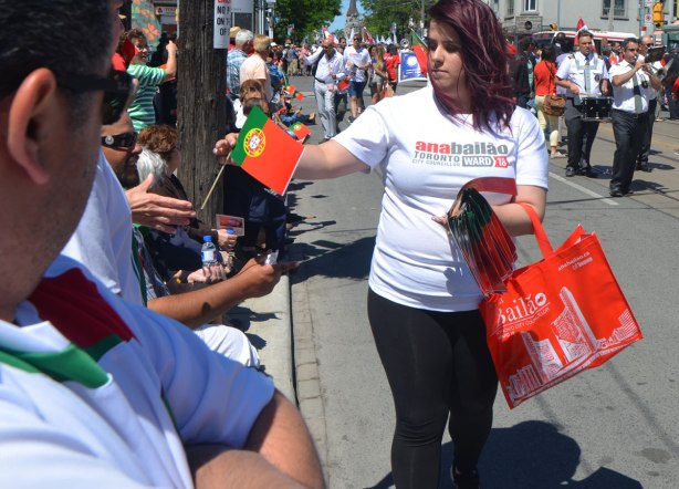 A woman is giving out small portuguese flags to people watching a parade. She is wearing a t-shirt in support of local politician Anna Bailao,